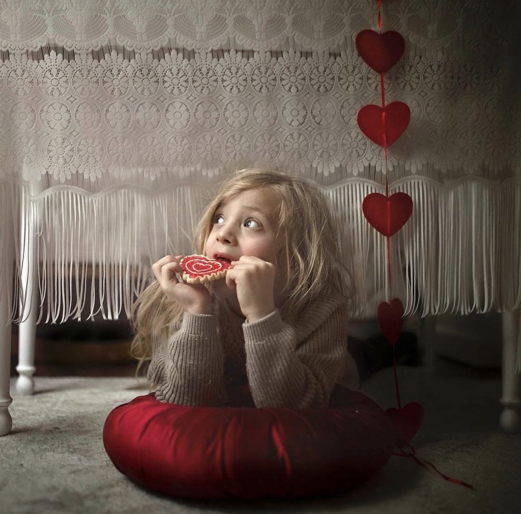 Valentines Day Photography. Fine art photograph of little boy eating heart shaped cookie under a table.