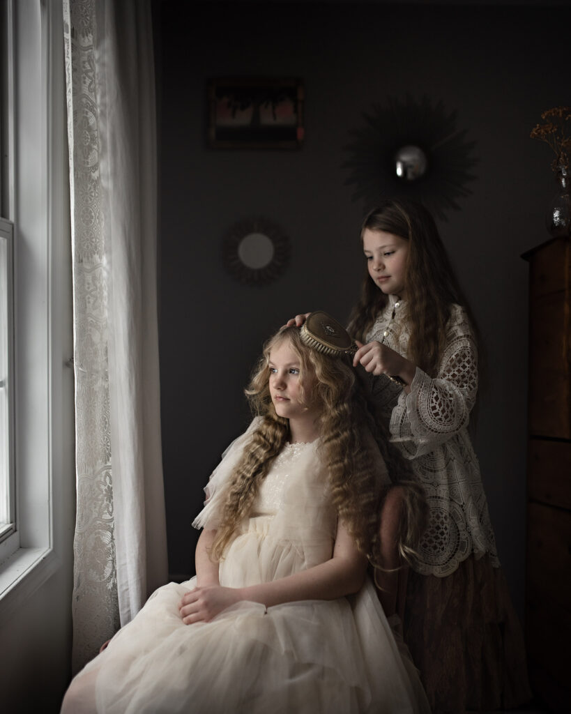 Fine Art Portrait Photograph. Sisters sitting at a Window. Younger sister brushing older sister's hair.